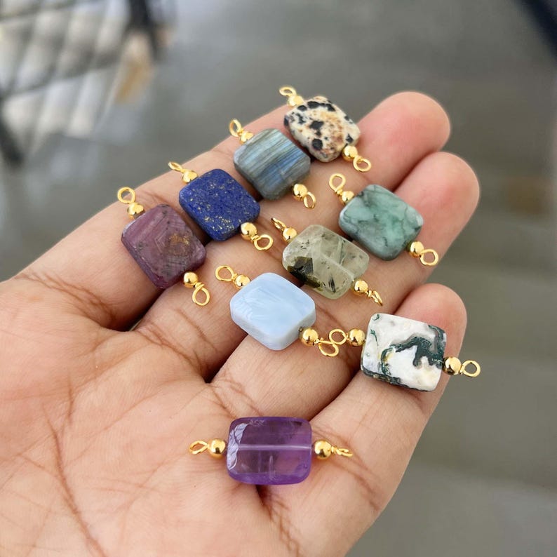 Hand holding a variety of natural crystal gemstone charms with gold bails. Square and rectangular pendants featuring amethyst, lapis lazuli, labradorite, dalmatian jasper, green moss agate, blue lace agate, phantom quartz, and white-green moss agate, ideal for handmade jewelry making.