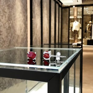 Op de afbeelding: Zilveren oorstekers, elk met een levendige rode edelsteen. De oorbellen worden gepresenteerd op een glazen vitrine, die het licht reflecteert. De achtergrond toont een winkelinterieur met mannequins.