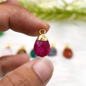 Op de afbeelding: Een druppelvormige hanger met een gefacetteerde, helder magenta edelsteen. De bovenkant van de hanger is afgewerkt met goudkleurig metaal en heeft een kleine lus. Andere hangers zijn zichtbaar op de achtergrond. Sieraden.