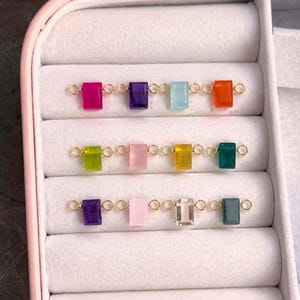 Puede incluir: Una colección de conectores de piedras preciosas de colores en varios tonos, incluyendo rosa, morado y verde, se muestra en una caja de joyería. Cada gema rectangular está conectada por pequeños anillos dorados. Las gemas están dispuestas en filas.