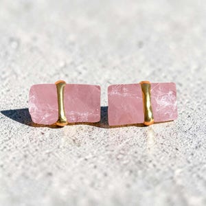 Op de afbeelding: Een paar rechthoekige rozenkwarts oorbellen. Elke oorbel heeft een lichtroze, ruw geslepen steen die op zijn plaats wordt gehouden door een goudkleurige metalen band. De oorbellen worden gepresenteerd op een lichtgrijze ondergrond.