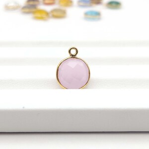 May include: A round, light pink gemstone pendant with a gold-colored metal frame. The gemstone has a faceted surface, and a small loop is at the top for attaching to a chain or other jewelry component. Other colorful pendants are visible in the background.