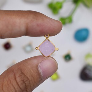 May include: A gold-colored pendant with a light purple, square-shaped gemstone. The gemstone is set in a gold frame with small decorative accents on each side. The pendant has a small loop at the top for a chain.