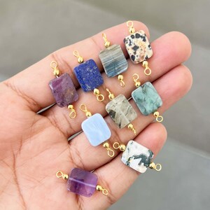 Hand holding a beautiful collection of natural crystal gemstone charms with gold bails. Square and rectangular pendants made from amethyst, lapis lazuli, labradorite, dalmatian jasper, green moss agate, blue lace agate, phantom quartz, and other semi-precious stones – perfect for jewelry making.