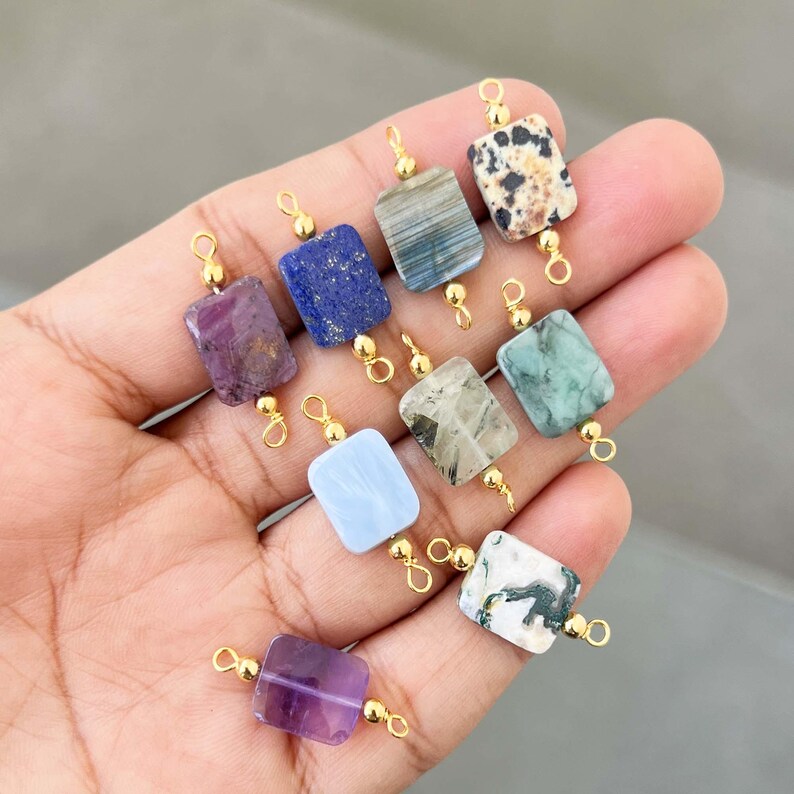 Hand holding a beautiful collection of natural crystal gemstone charms with gold bails. Square and rectangular pendants made from amethyst, lapis lazuli, labradorite, dalmatian jasper, green moss agate, blue lace agate, phantom quartz, and other semi-precious stones – perfect for jewelry making.
