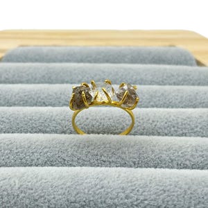 May include: A gold-colored ring featuring three raw, uncut gemstones. The gemstones are a mix of clear and smoky brown hues, set in a simple, elegant design. The ring is displayed on a gray, textured surface.