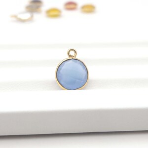 May include: A round, light blue gemstone pendant with a gold-colored bezel. The pendant has a faceted surface and a small loop at the top for attaching to a chain or bracelet. Other colorful pendants are slightly visible in the background.