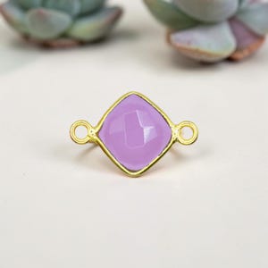 May include: A gold-colored metal connector with a light purple, faceted, square-shaped center. The connector has two small loops on either side. The background is a soft, neutral color with blurred green plants.