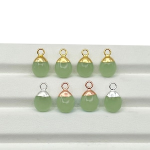 May include: Eight teardrop-shaped pendants with a light green, oval-shaped stone. The pendants have gold or silver-colored metal caps and loops. The pendants are displayed on a white surface.