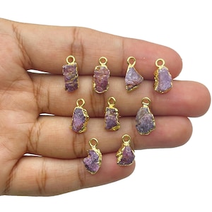 May include: A selection of raw, purple gemstone pendants with gold-coloured edges and small loops for hanging. The pendants vary in shape and size, showcasing the natural texture and colour variations of the stones.