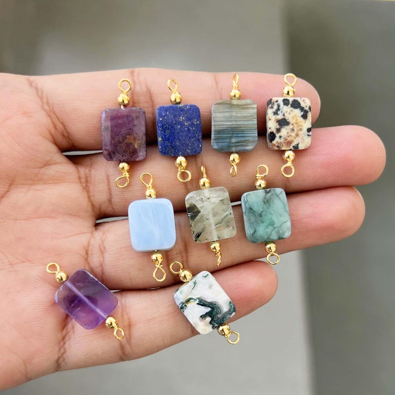 Hand holding a collection of natural crystal gemstone charms with gold bails. Includes square and rectangular pendants made of amethyst, lapis lazuli, blue lace agate, moss agate, dalmatian jasper, and other semi-precious stones, perfect for jewelry making