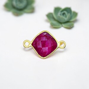 May include: A gold-toned jewelry component with a faceted, square-shaped, magenta-colored gemstone. The component has small loops on either side. Two green succulents are in the background on a white surface.