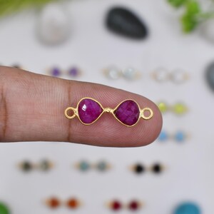 Könnte beinhalten: Ein goldfarbenes Schmuckelement mit zwei fuchsiafarbenen, tropfenförmigen Steinen. Die Steine sind durch einen goldfarbenen Rahmen mit kleinen Schlaufen an jedem Ende verbunden. Das Element wird von einem Finger gehalten.
