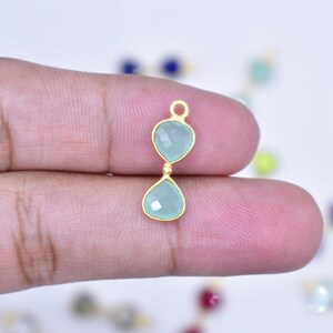 May include: A gold-toned pendant featuring two faceted, pear-shaped light green stones arranged vertically. The stones are set in gold-toned metal bezels, with a small circular jump ring at the top for attachment to a chain or jewelry finding.