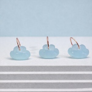 May include: Three pairs of cloud-shaped earrings. Each earring has a light blue, translucent cloud charm hanging from a rose gold hoop. The earrings are displayed on a white surface against a light blue backdrop.