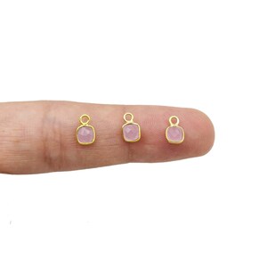 May include: Three small, square-shaped pink gemstone charms with gold-colored frames. Each charm has a loop at the top for attaching to jewelry. The charms are displayed against a light background.