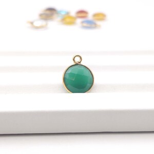 May include: A faceted, round, green gemstone pendant with a gold-colored bezel and a small loop at the top. Other colorful pendants are slightly out of focus in the background. The pendant is displayed on a white surface.