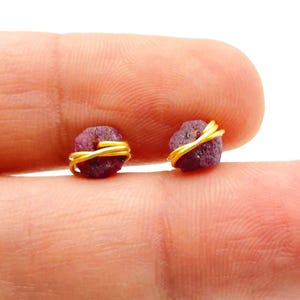May include: Two small, rough-cut ruby gemstones are wrapped with gold-coloured wire. The gemstones are a deep reddish-purple colour and are displayed against a neutral background. These earrings are a unique jewellery item.