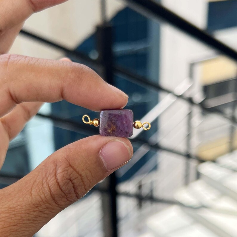 Close-up of fingers holding a single natural ruby in zoisite gemstone charm with gold bail. The square purple pendant shows deep magenta color with golden and black inclusions, perfect for handmade jewelry making.
Hand holding a natural ruby in zoisite square gemstone pendant with gold bail.
Natural Ruby in Zoisite Slice Charm with Gold Bail – Close-up of a vibrant purple ruby in zoisite rectangular pendant with golden inclusions, held between fingers for jewelry making.