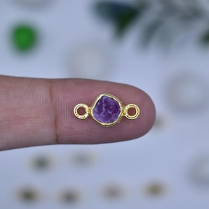 May include: A gold-colored connector with a raw, purple gemstone center. The gemstone is irregularly shaped and set within a gold bezel. The connector has small loops on either side for attaching to other jewelry components.