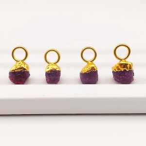 Peut inclure: Quatre pendentifs en pierres précieuses plaqués or, de forme brute et organique. Chaque pendentif présente une pierre brute de couleur violet foncé, surmontée d'une boucle dorée texturée pour la suspension. Les pendentifs sont présentés sur une surface blanche.