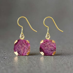 May include: A pair of drop earrings featuring rough-cut, deep magenta-coloured stones set in gold-toned metal prongs. Each stone has a square-like shape with textured surfaces, suspended from simple gold-toned curved ear wires for a dangling effect.