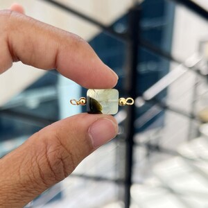 Close-up of fingers holding a single natural prehnite gemstone charm with gold bail. The square pendant displays a beautiful pale green color with dark inclusions, ideal for handmade jewelry making.
Natural Prehnite Slice Charm with Gold Bail – Close-up of a polished pale green prehnite rectangular pendant with dark inclusions, held between fingers for jewelry making.