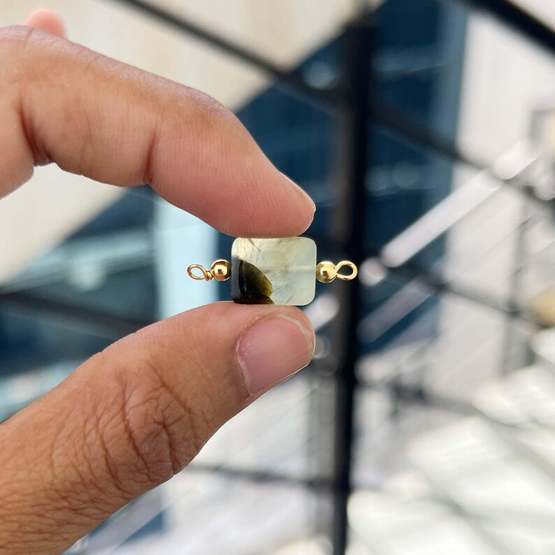 Close-up of fingers holding a single natural prehnite gemstone charm with gold bail. The square pendant displays a beautiful pale green color with dark inclusions, ideal for handmade jewelry making.
Natural Prehnite Slice Charm with Gold Bail – Close-up of a polished pale green prehnite rectangular pendant with dark inclusions, held between fingers for jewelry making.