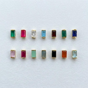 May include: A collection of rectangular gemstone studs in various colors, including green, pink, red, and blue, set in gold. The gemstones are arranged in two rows against a white background, showcasing their vibrant hues and elegant design.