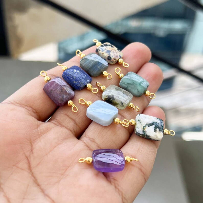 Hand holding a beautiful collection of natural crystal gemstone charms with gold bails. Square and rectangular pendants made from amethyst, lapis lazuli, labradorite, dalmatian jasper, green moss agate, blue lace agate, phantom quartz, and other semi-precious stones – perfect for jewelry making.