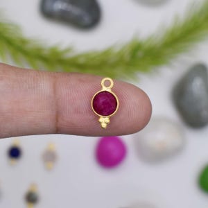 May include: A small, gold-coloured pendant featuring a round, deep red gemstone. The pendant has a loop at the top and three small decorative beads at the bottom. The gemstone is set in a gold bezel.