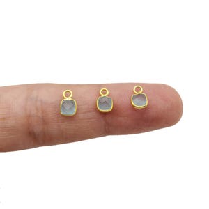 May include: Three small, gold-colored pendants with square, light blue gemstones. Each pendant has a small loop at the top for attaching to a chain or other jewelry component. The pendants are displayed on a finger.