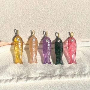 May include: Five fish-shaped pendants in various colours. The pendants are yellow, peach, purple, dark green, and pink. Each has a small, clear gem and a gold-coloured loop at the top for hanging.