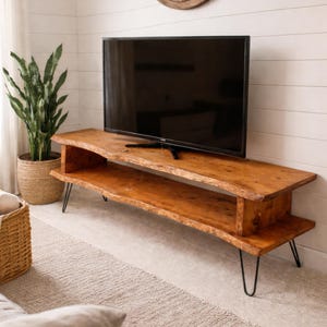 May include: A natural wood TV stand with a live edge design. The stand has a lower shelf and is supported by black hairpin legs. A large flat-screen TV sits on top. A woven basket and a potted plant are visible.