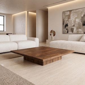 May include: A low-profile square coffee table crafted from dark brown wood with a visible grain pattern. The table features a thick, flat tabletop surface supported by a central block base, positioned centrally in a room with white sofas.
