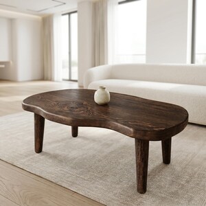 May include: A dark brown wooden coffee table with an irregular, organic kidney-bean shape and four sturdy, tapered legs. The surface displays a visible wood grain pattern and a smooth finish. A small, off-white ceramic vase sits on the table.