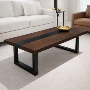 Peut inclure: Une table basse rectangulaire avec une finition en bois foncé et une bande noire au centre. La table a des pieds en métal noir. Un bol doré est posé sur la table. La table est dans un salon.
