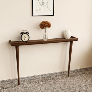 May include: A dark brown console table in a mid-century modern style, featuring a rectangular top and four tapered legs. It's adorned with a black alarm clock, a clear vase with dried flowers, and a white geometric vase. A framed line drawing is above.