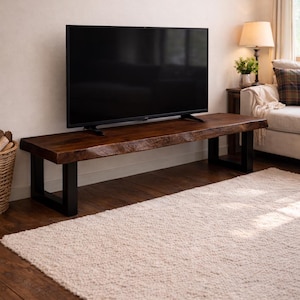 May include: A dark brown, live-edge TV stand with black metal legs supports a large black television. The stand sits on a light-coloured, textured rug in a living room setting. A lamp and sofa are visible in the background.