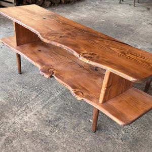 May include: A handcrafted wooden coffee table with a natural edge design. The table features two tiers, supported by four slender legs. The wood has a warm, reddish-brown stain, showcasing the natural grain and knots. A rustic, yet modern piece of furniture.