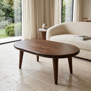 May include: A low oval coffee table crafted from dark brown wood with a smooth, polished surface. The table features four tapered, cylindrical legs that extend outward from the base, providing a stable foundation for this piece of furniture.