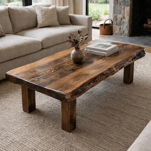 May include: A dark wood coffee table with a live edge, approximately 150cm long, in a living room. The table has a rectangular shape and sits on four square legs. A vase with dried flowers and books are on the table. The room has a neutral colour scheme.