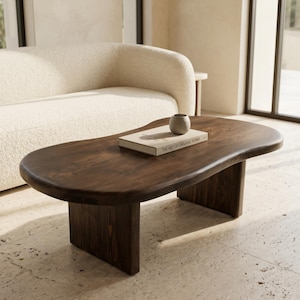 May include: A dark brown wooden coffee table with an irregular, organic-shaped top and two thick, rectangular slab legs. A light-colored book titled The Art of Organic Form and a small, plain gray ceramic bowl sit on the table surface.