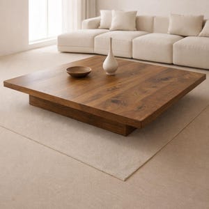 May include: A low, square wooden coffee table with a rich, warm wood grain finish. A small, round wooden bowl and a white, teardrop-shaped vase sit on the table. The table is set on a neutral-toned rug, with a cream-colored sectional sofa in the background.