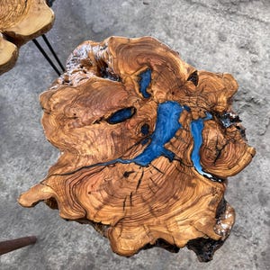 May include: A wooden table with a distinctive, organic form. The table showcases a rich, brown wood grain, complemented by bright blue resin accents that fill the natural cracks. It is supported by black metal hairpin legs. The table's dimensions are in centimetres.