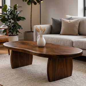 May include: A dark brown, oval-shaped wooden coffee table with two curved, vertical supports. The table sits on a light beige rug in a living room setting. A white vase with dried wheat stalks sits on the table.