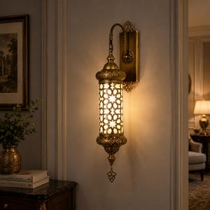 Puede incluir: Un aplique de pared con acabado en latón antiguo y cuerpo cilíndrico con intrincados recortes geométricos en forma de estrella. La lámpara emite un brillo cálido a través del metal calado y cuenta con un remate decorativo en la base.