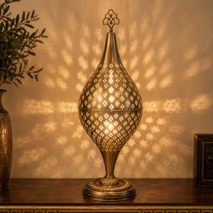May include: A teardrop-shaped metal table lamp with an antique brass finish, featuring intricate laser-cut geometric patterns that cast star-shaped light projections onto the wall. The lamp has a pointed finial top and a circular base.