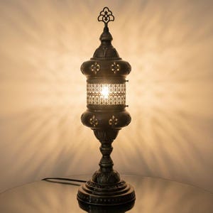 Könnte beinhalten: Eine verzierte Tischlampe in Bronzefarbe mit dekorativem Filigran-Schirm. Die Lampe hat einen detaillierten Sockel und eine gemusterte obere Spitze. Das Licht scheint durch die aufwendigen Ausschnitte und erzeugt einen warmen Schein. Die Lampe ist etwa 46 cm hoch.