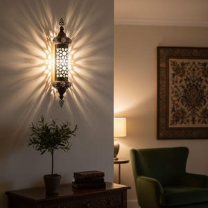 Könnte beinhalten: Verzierte Wandleuchte aus Bronze mit Sternmuster, die ein strahlendes Licht wirft. Die Leuchte ist an einer neutralfarbenen Wand angebracht und erzeugt eine warme Atmosphäre. Die Leuchte ist etwa 61 cm hoch.